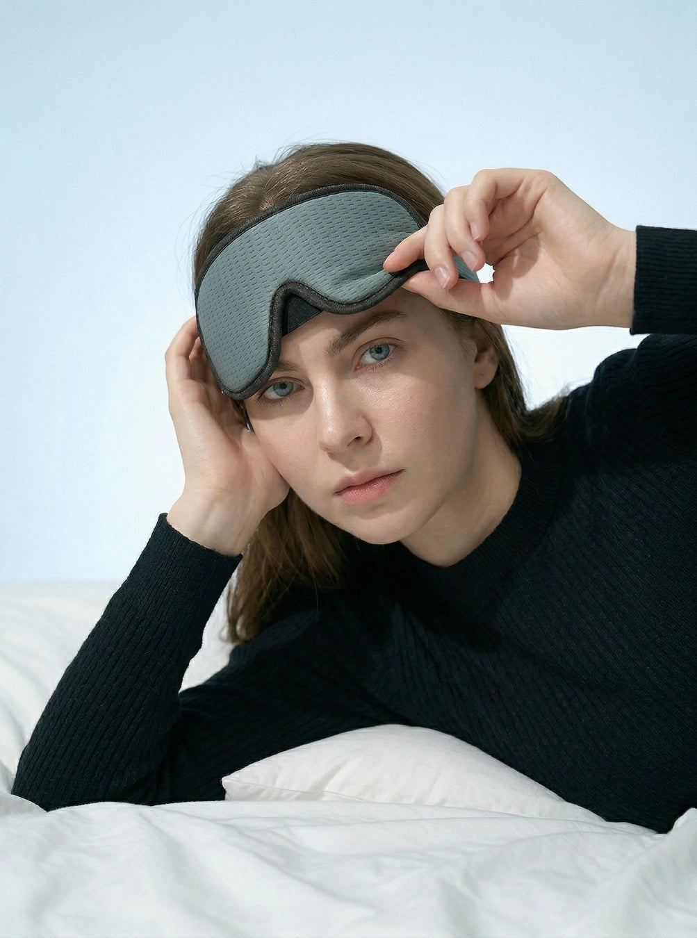 Person wearing a gray sleep mask in bed with a light blue wall background, dusk