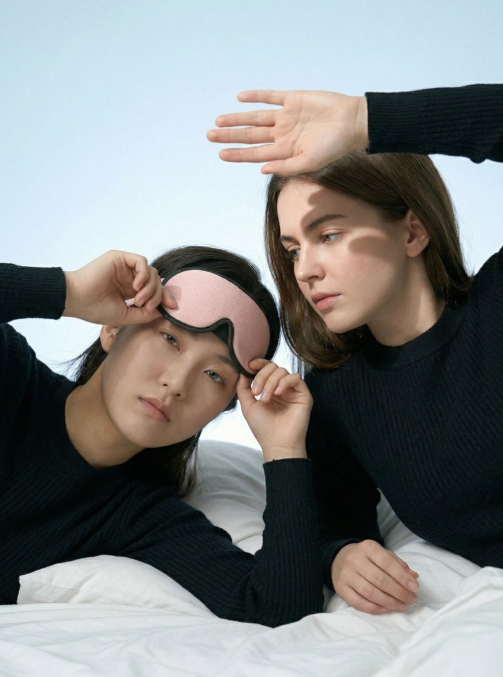 Two people in bed with one wearing a pink sleep mask and the other adjusting it, peach