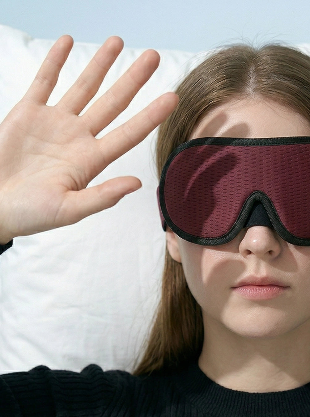 Person wearing a sleep mask with one hand raised in front of their face, maroon
